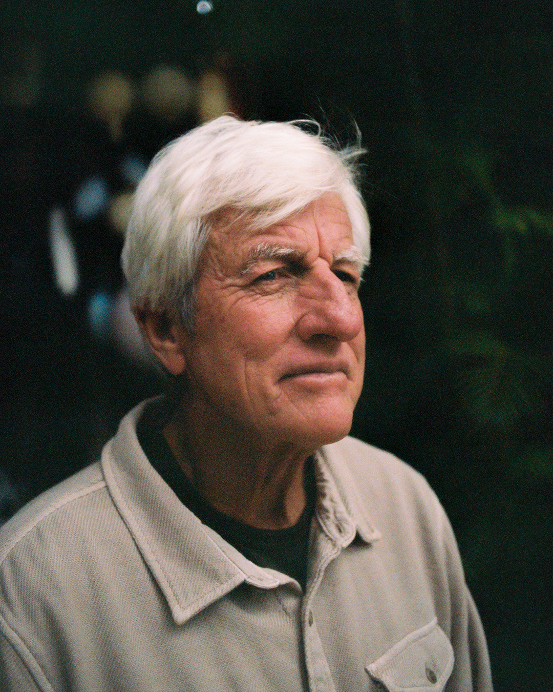 An elderly man with white hair, wearing a beige jacket over a black shirt, looking thoughtfully into the distance outdoors.
