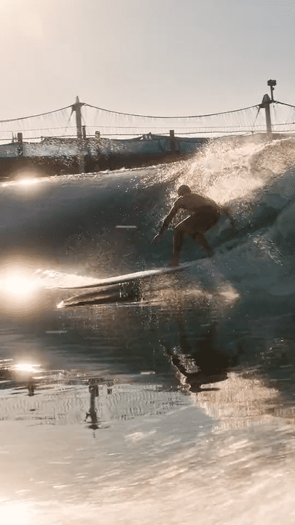 A person surfing on a wave during sunset at the beach with a pier and some structures in the background.