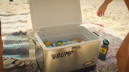 An open cooler filled with drinks on a beach with towels and a person's arm visible.