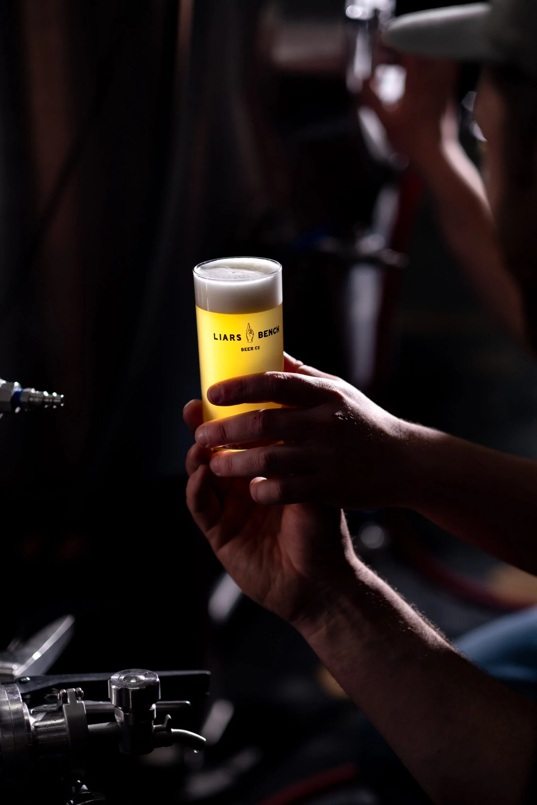 A person holding a glass of beer with a foamy head, with the beer illuminated by a light, in a dark bar environment. The glass has 'Liars Bench Beer Co' printed on it.