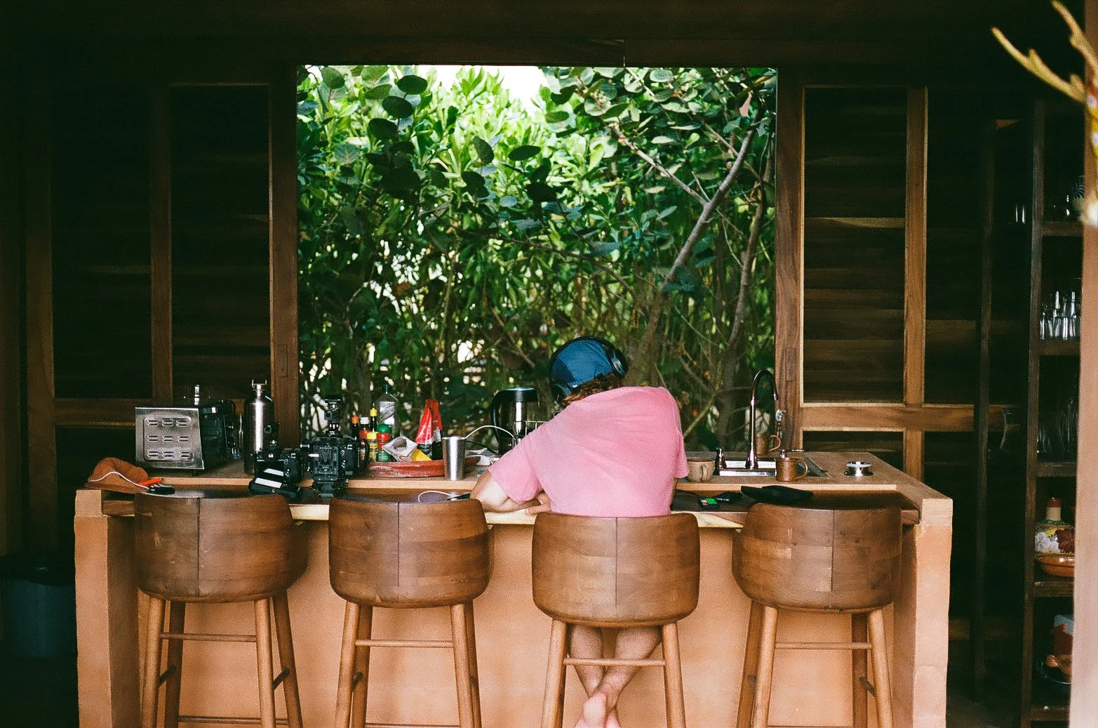 A person sitting at a wooden kitchen or bar counter, wearing a pink shirt and a blue helmet, is looking down at the counter. The background shows lush green foliage outside a large window, with the room's interior featuring wooden walls and shelves with glasses. The counter has various kitchen items and utensils.