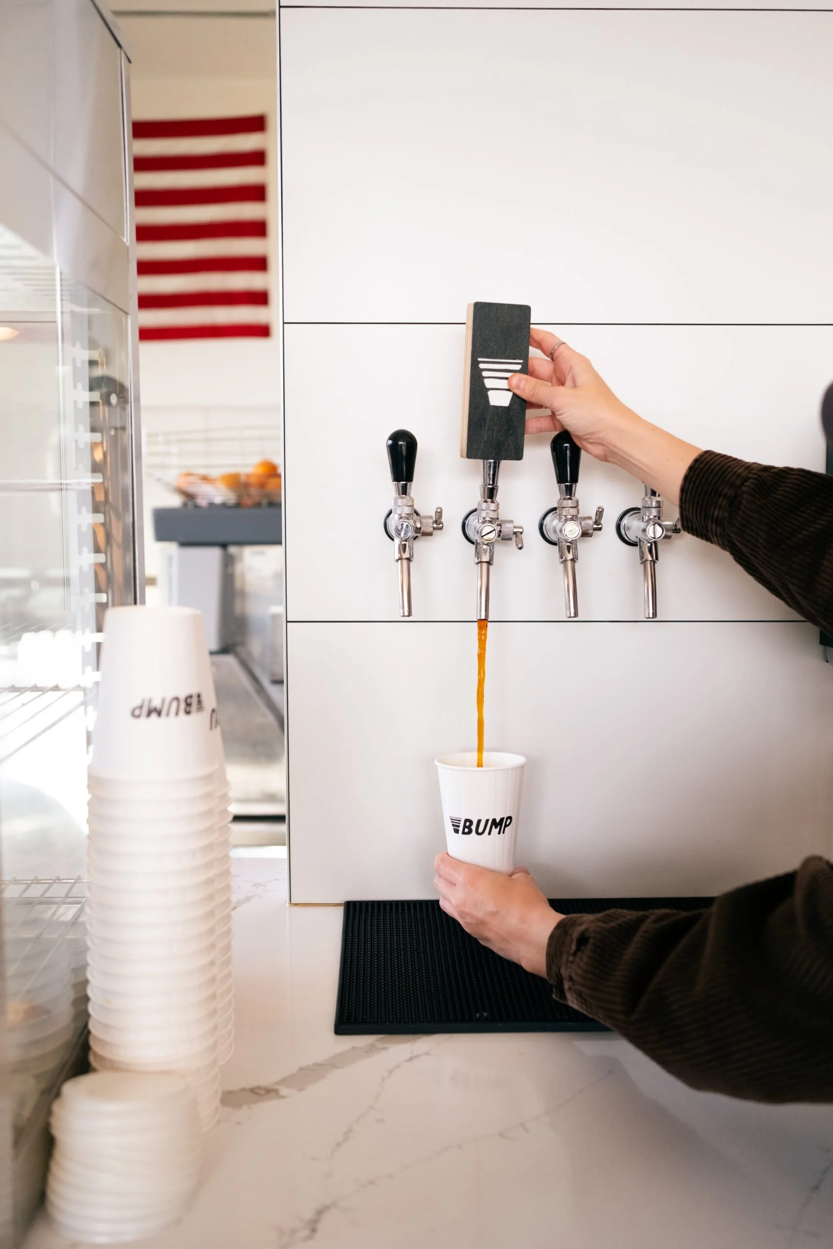 A person is filling a white cup with a brown beverage from a tap at a cafe, with several other taps above. To the left, a stack of white cups with 'BUMP' printed on them is visible.