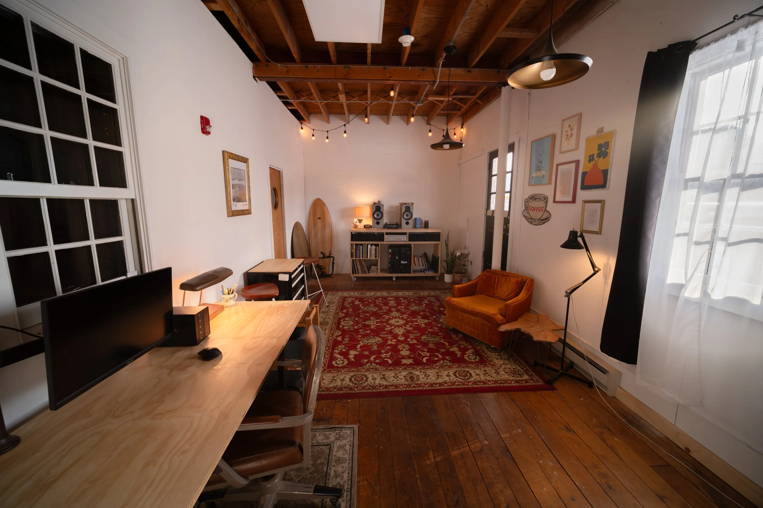 A cozy living room with a wooden floor, white walls, and a high wooden ceiling. There is a large window with white curtains on the right, a red patterned rug on the floor, and a small orange velvet sofa. In the back, a sound system with speakers and a stereo on a white console table. On the left, a workspace with a long wooden desk, a computer monitor, and chairs. Decor includes framed artwork and surfing boards leaning against the wall.