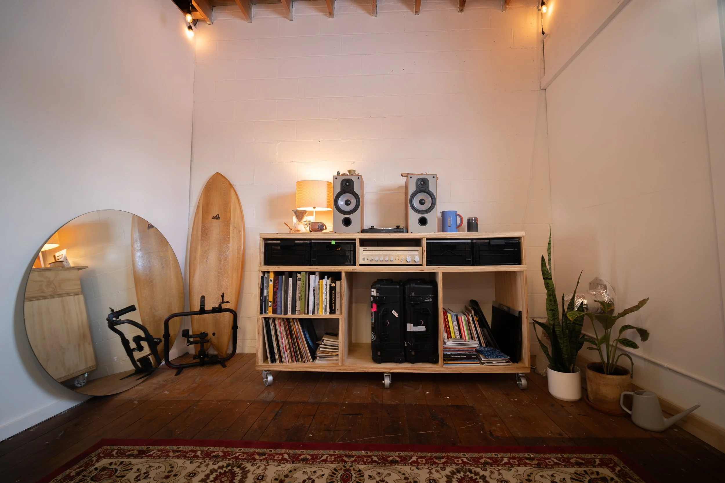 A cozy room corner with a wooden shelving unit holding books, vinyl records, and audio equipment. Two large speakers sit on top of the shelf. To the left, round mirrors, skateboards, and exercise equipment lean against the wall. To the right, there's a potted plant, watering can, and a lamp with a shade, with soft lighting and a hardwood floor.