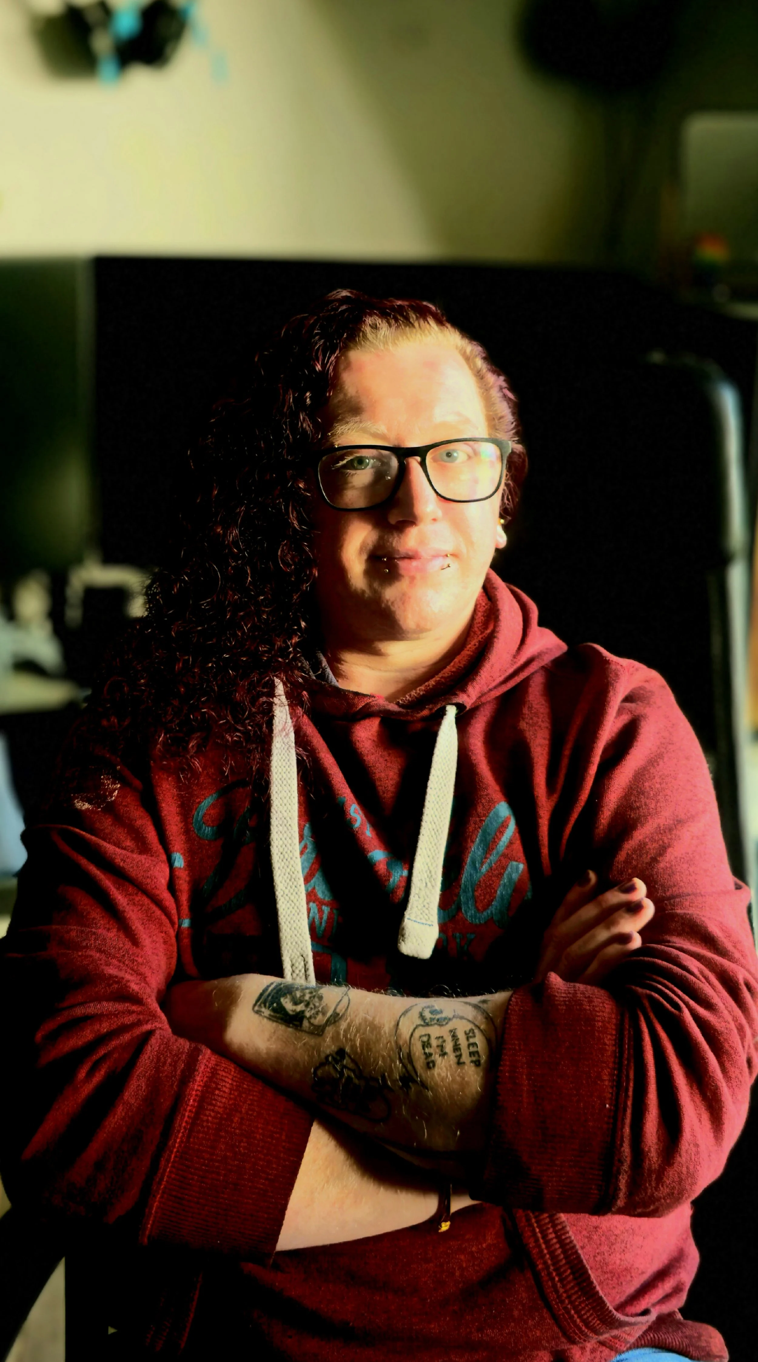 Person with curly hair and glasses sitting with arms crossed, wearing a red hoodie, in a room with a dark background and shadows.