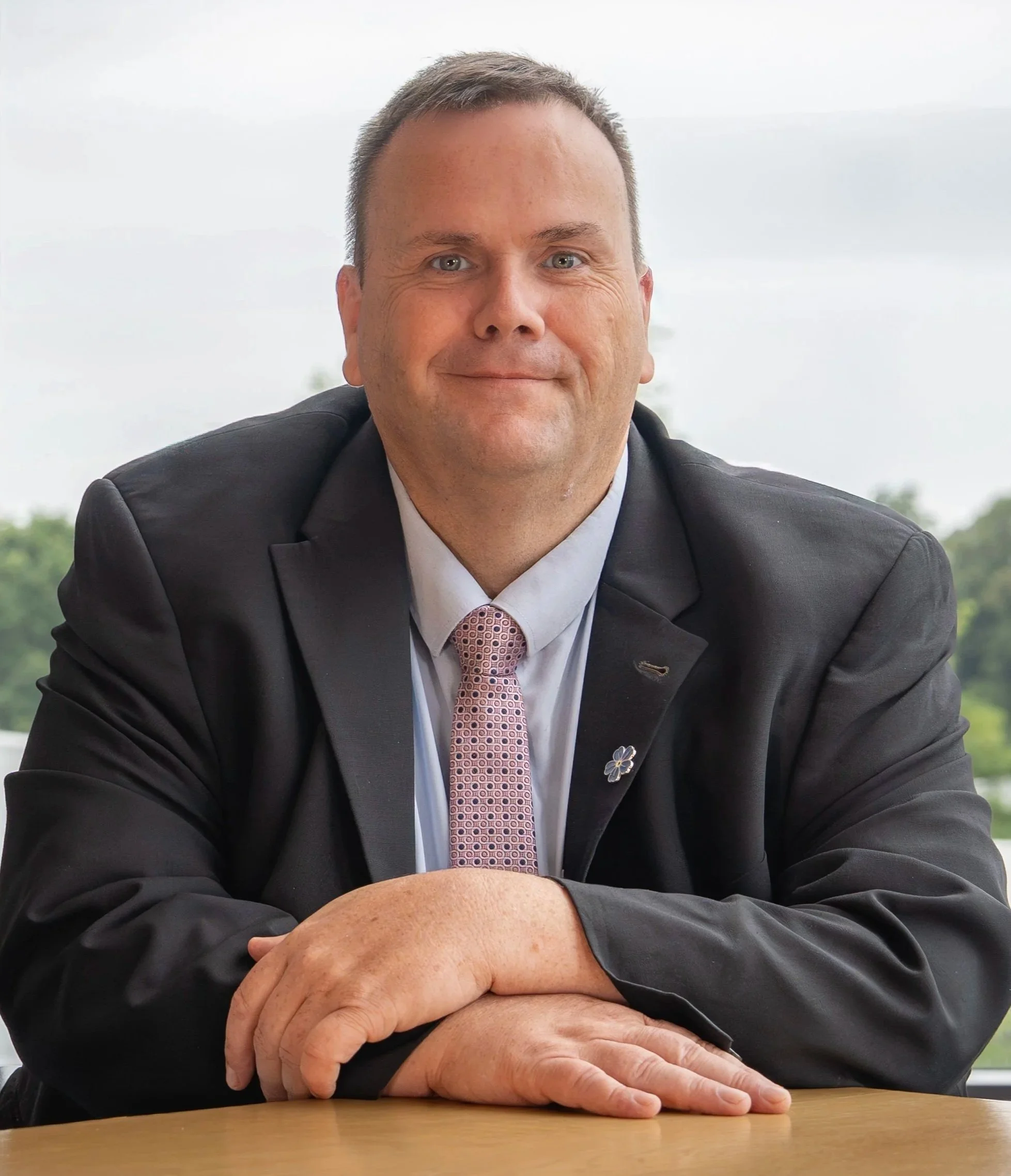 A man in a black suit and light pink patterned tie sitting at a wooden table with arms crossed, smiling, with an outdoor background.