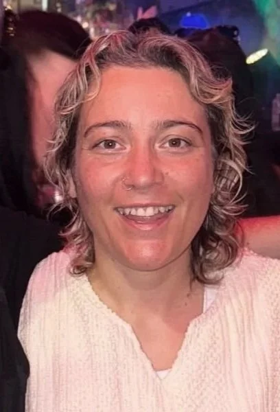 A woman with curly, shoulder-length hair and a nose ring, smiling at a social event with colorful lights in the background.