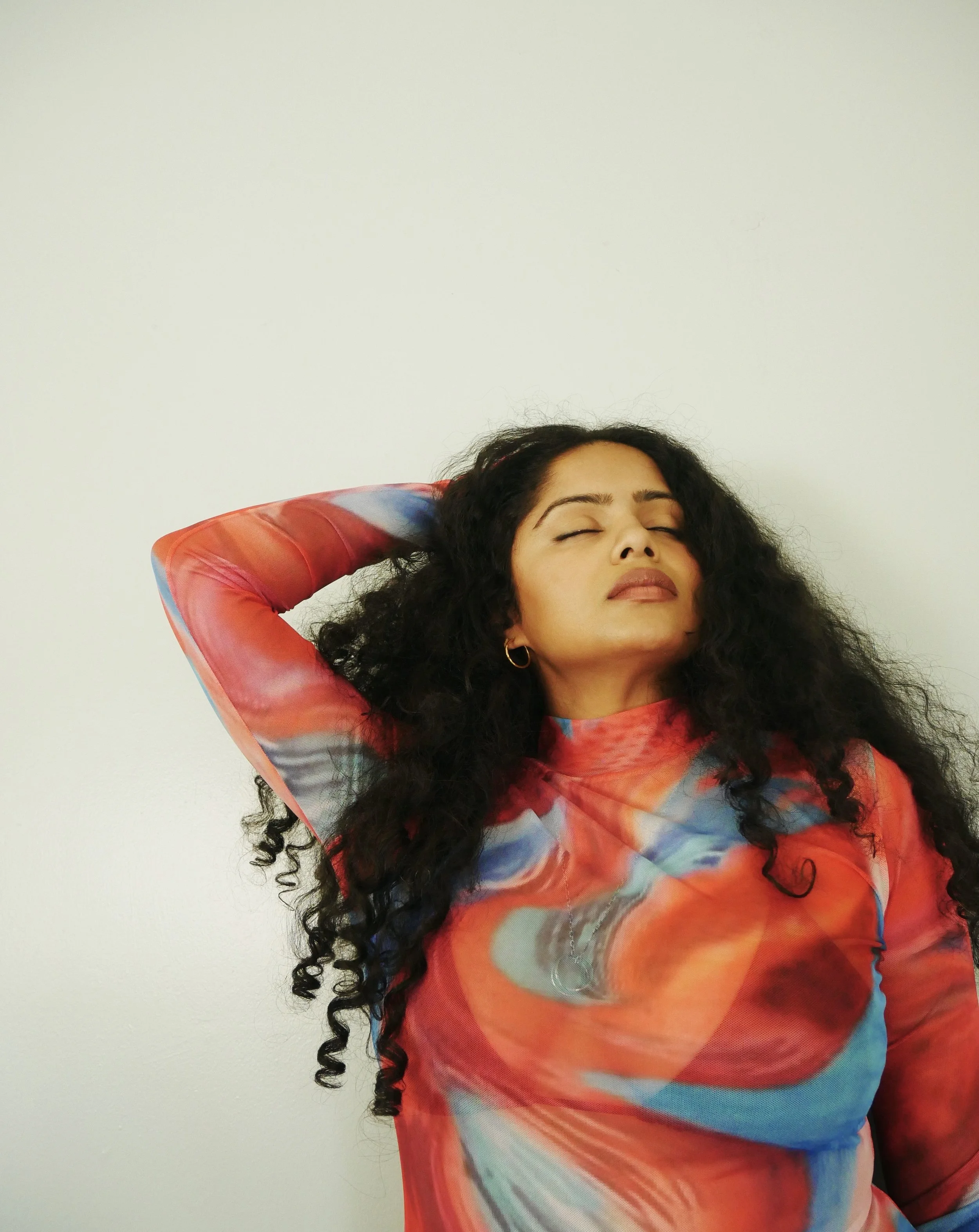 Woman with long curly black hair and hoop earrings, wearing a colorful abstract-patterned top, with eyes closed and hand resting on the back of her head, standing against a plain white wall.