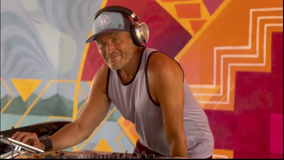 A man with a beard, wearing a baseball cap, headphones, and a sleeveless shirt, is DJing in front of a colorful, abstract mural.