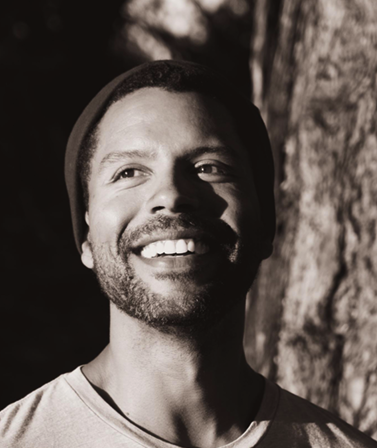 A smiling man outdoors near a tree, wearing a beanie and a light-colored shirt.