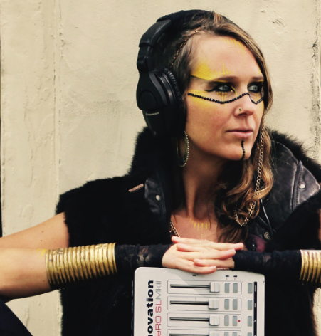 Woman wearing headphones with artistic makeup, leather vest, and metallic arm accessories, leaning on a keyboard synthesizer against a plain wall.