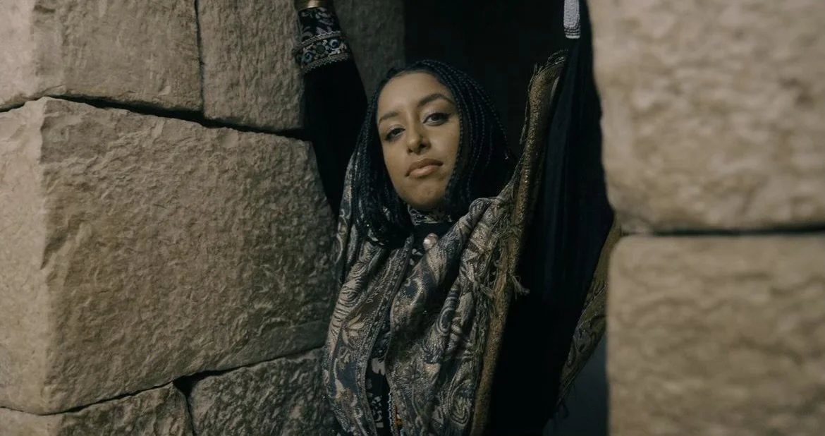 Egyptian American Woman with braided hair and patterned scarf looking through stone walls.