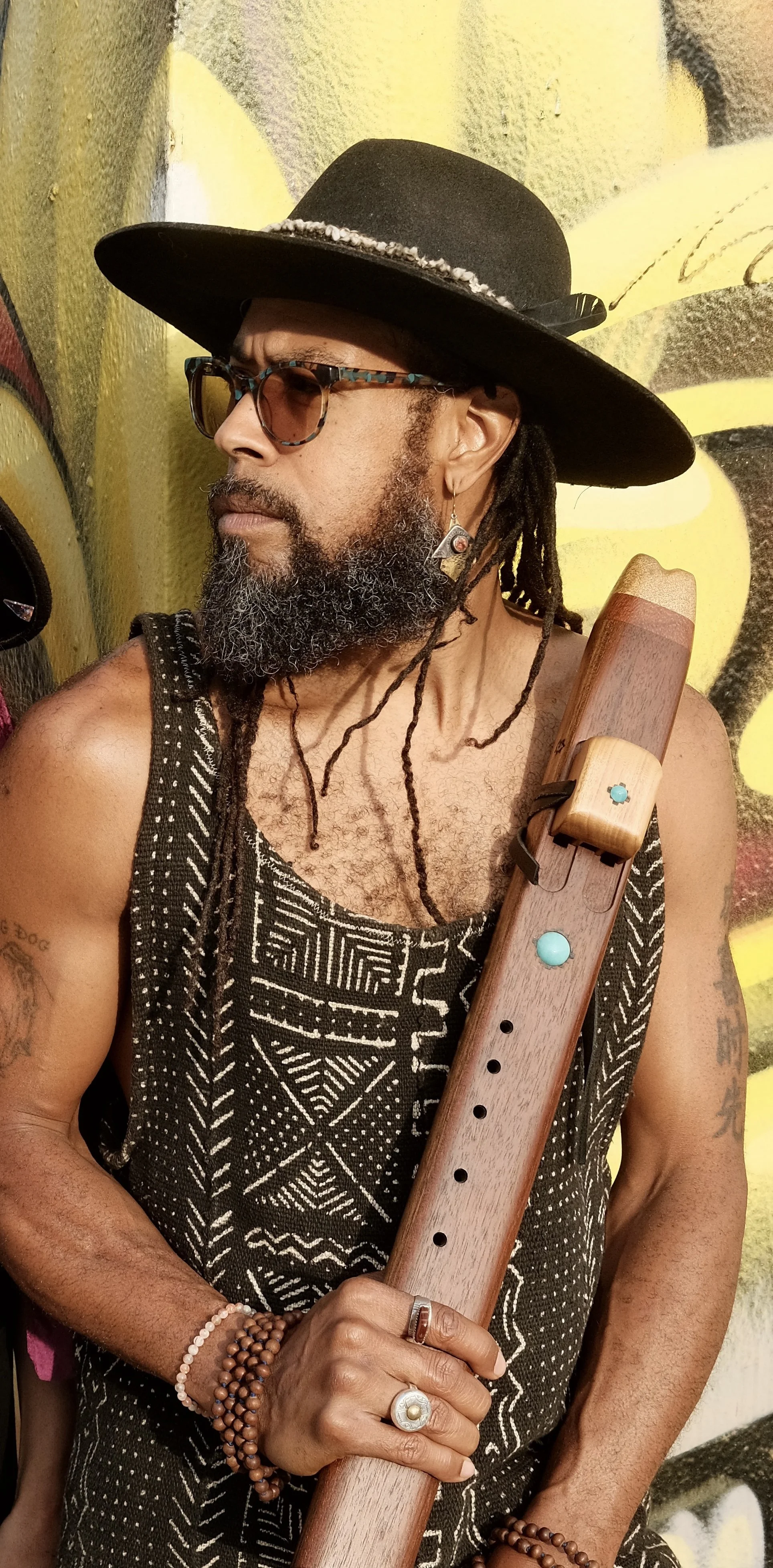 A man with a beard and dreadlocks wearing a black wide-brim hat, sunglasses, and a patterned black sleeveless shirt, holding a wooden flute, standing against a graffiti wall.