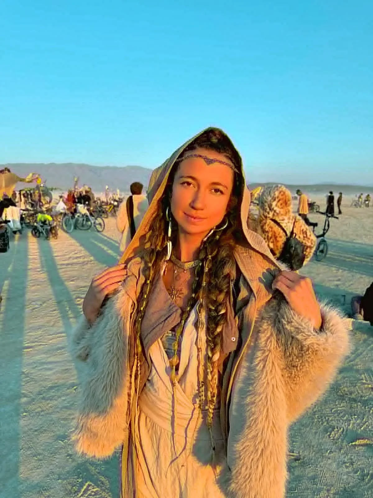 A young woman with braided hair, wearing a fur-lined jacket and a hood, standing on a desert at sunset with people and bicycles in the background.
