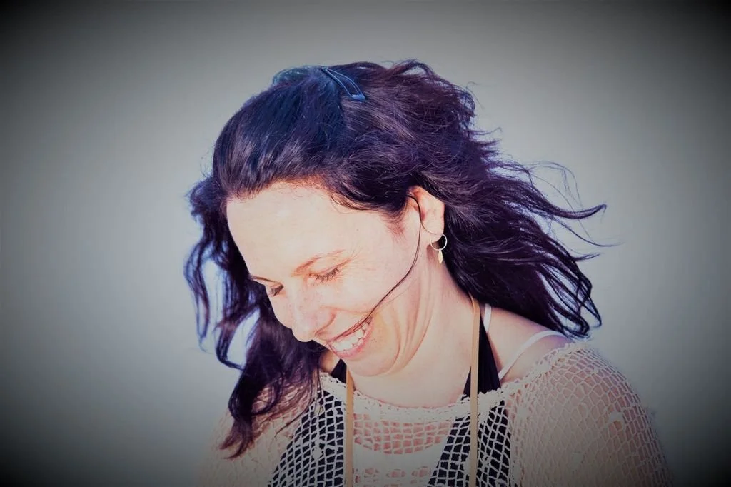 A woman with long dark wavy hair smiling and looking downward, wearing earrings, a netted top, and glasses hanging around her neck.