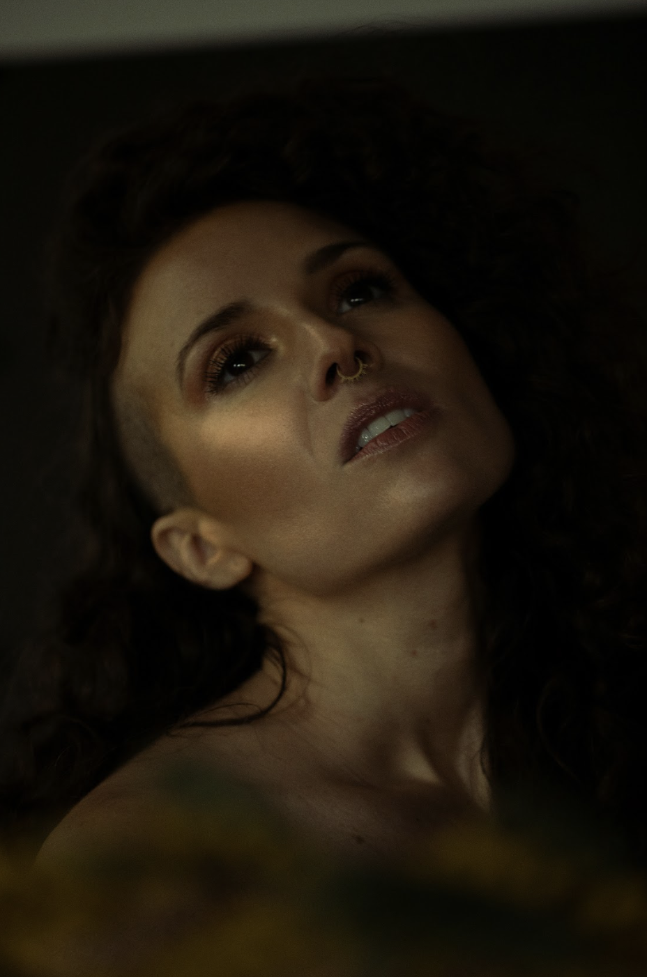 A woman with curly dark hair, wearing makeup and a septum piercing, lying down with her head tilted back, looking upward.