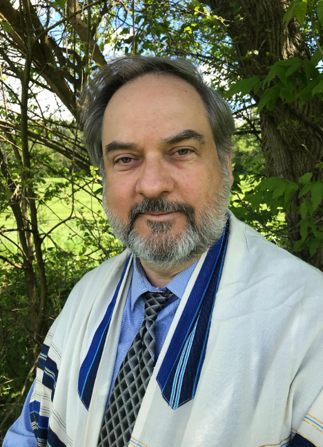 A man with a beard and mustache wearing a light-colored religious or academic robe over a blue shirt and a patterned tie, standing outdoors among green foliage and trees.