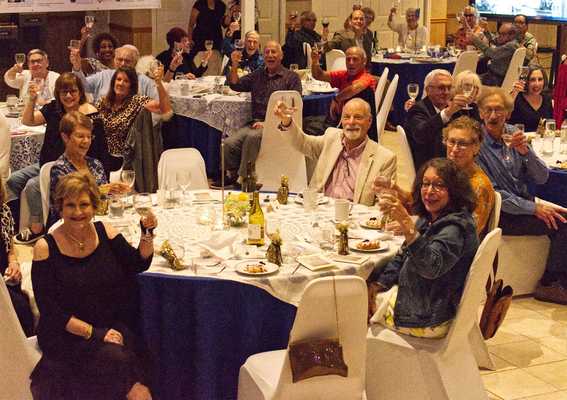 A group of people at a celebration dinner raising glasses of wine in a toast, seated around decorated tables at an indoor event.