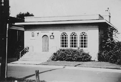 Black and white photo of a house with three arched windows, a wooden door, and steps leading up to the entrance surrounded by bushes.
