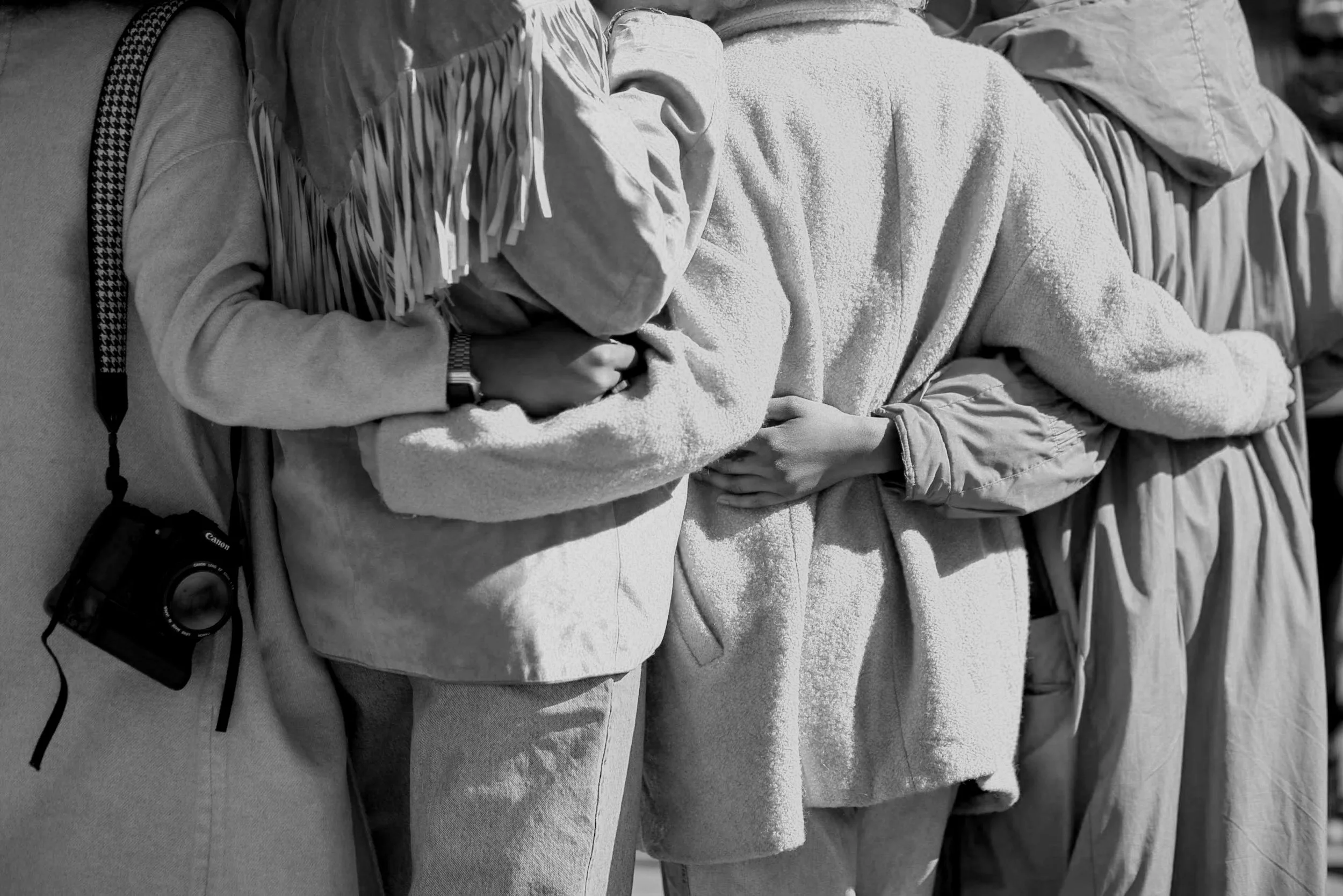 A group of people standing together, with some holding hands, wearing jackets and casual clothing. One person has a camera hanging from their shoulder.