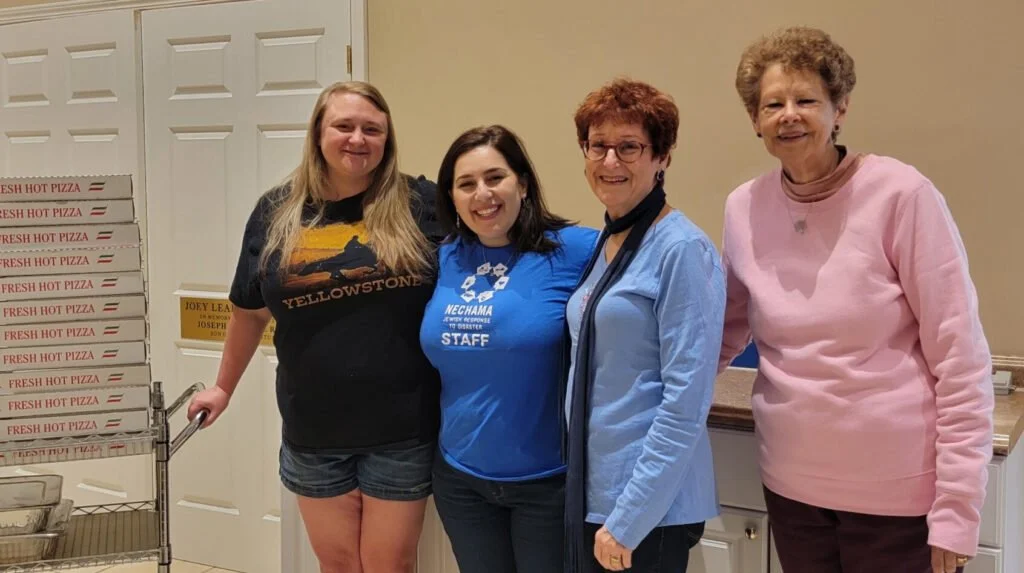 Four women standing side by side in a room, smiling. Behind them are stacked pizza boxes labeled "FRESH HOT PIZZA" and a pizza cart. The women are dressed casually.