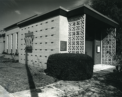 A mid-century modern building with patterned concrete blocks near the entrance, a small bush in front, and a large palm sculpture on the wall, in black and white.