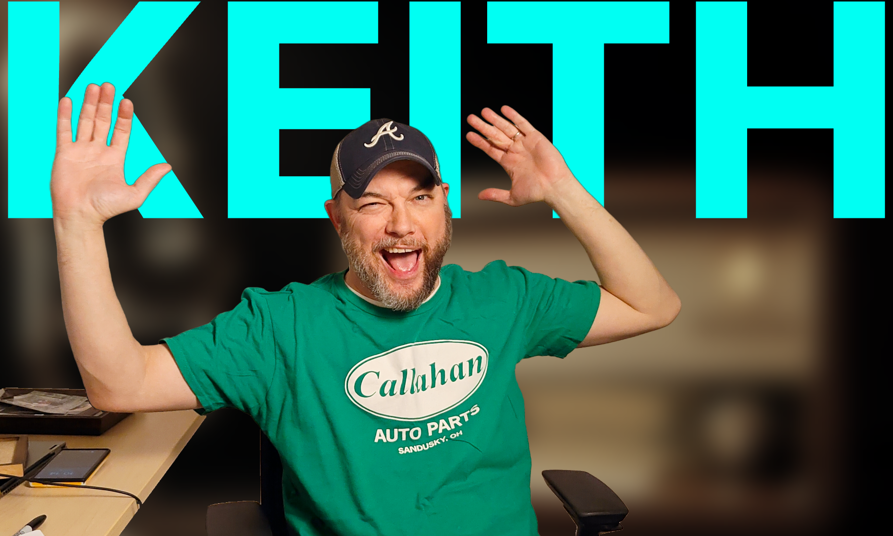 A man wearing a green t-shirt with 'Calahan AUTO PARTS' printed on it, a black baseball cap with an 'A' logo, winks and wags his tongue with a joyful expression, while raising both hands near his head. Behind him is a large sign that reads 'KEITH' in bright blue letters.