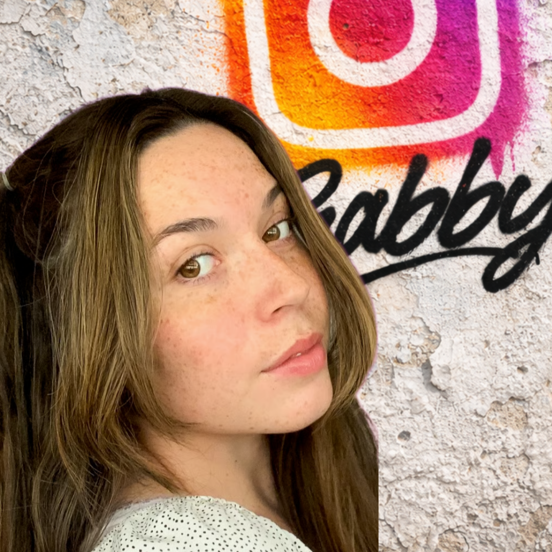 A young woman with light skin and brown hair with highlights, looking over her shoulder at camera, standing in front of a colorful graffiti wall with the words 'Baby' and a stylized logo.