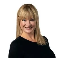 Portrait of a woman with blonde hair and bangs, wearing a black top, smiling against a plain white background.