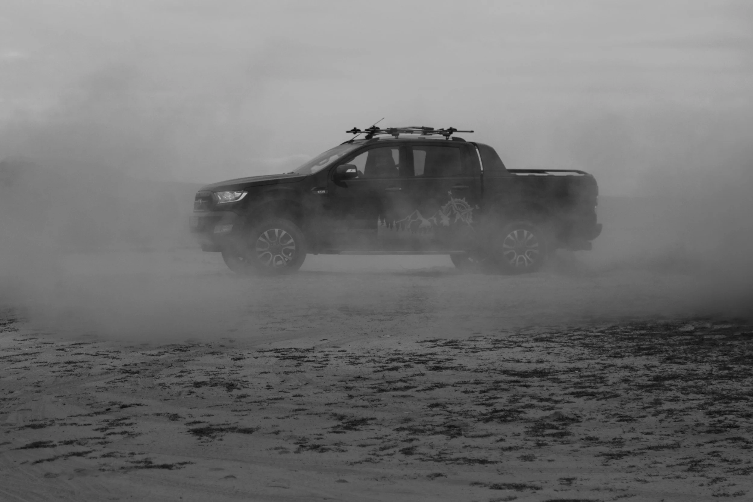 Black pickup truck driving through a dusty or sandy environment with dust cloud around it, under cloudy sky, in black and white