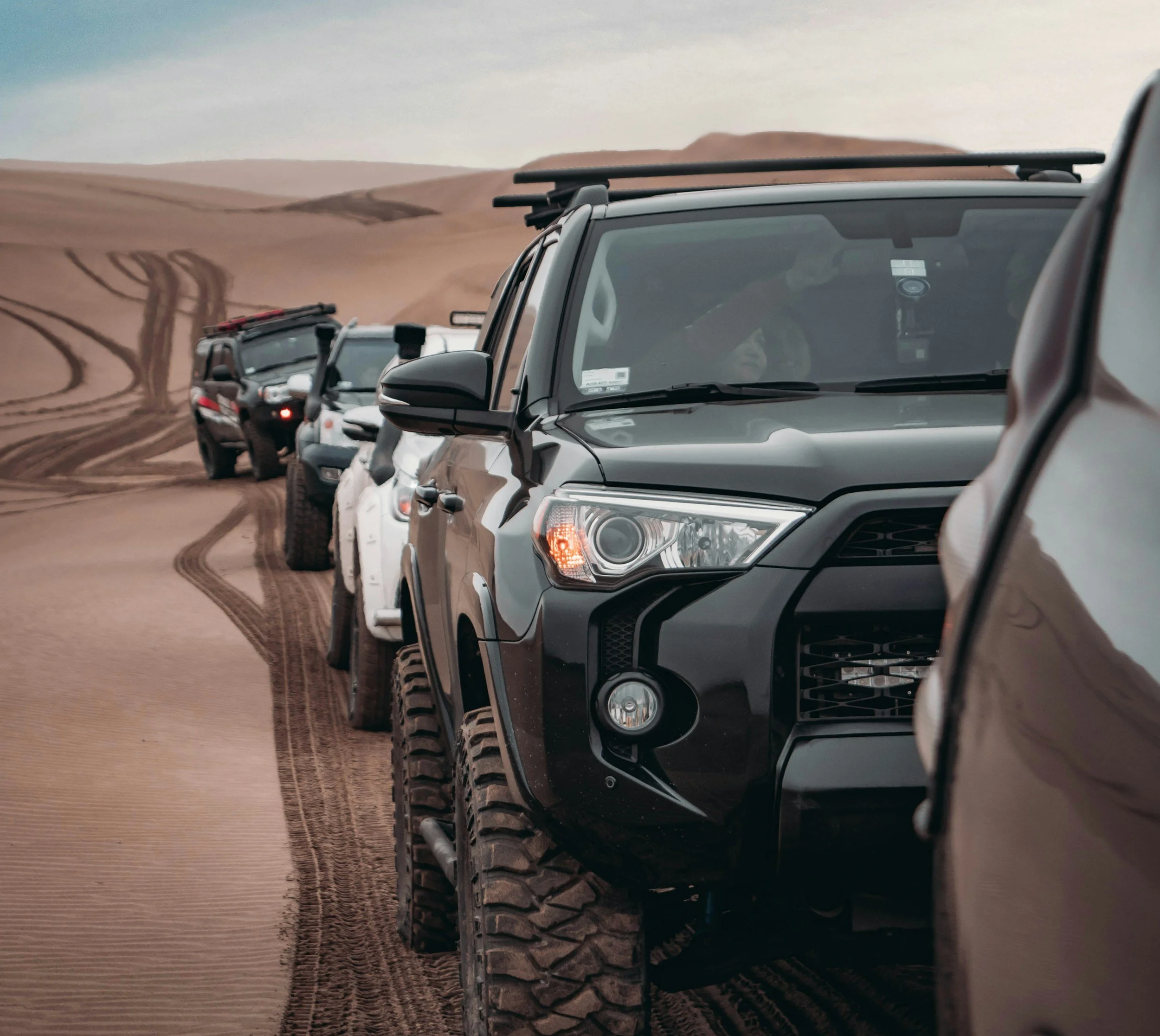 Line of 4 off-road vehicles driving on sand dunes with tire tracks visible on the sand.