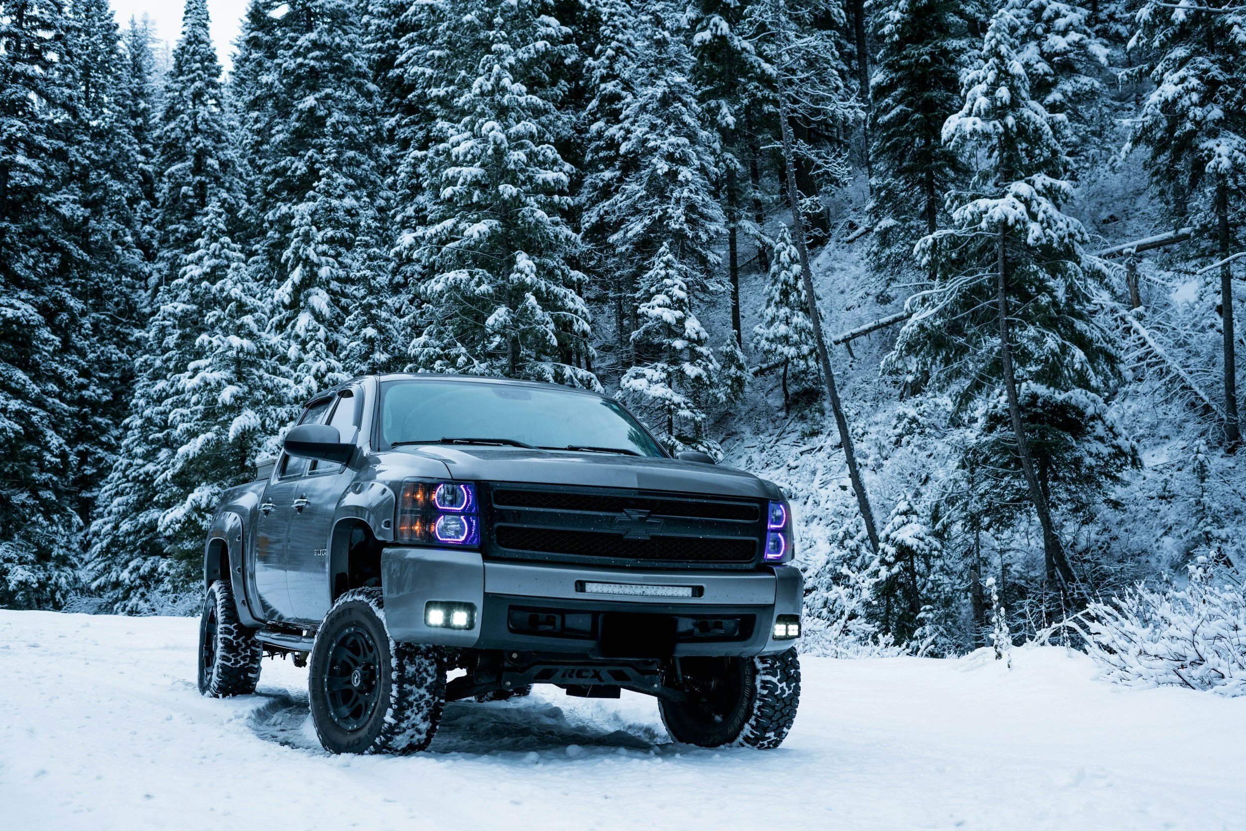 A silver pickup truck with off-road tires parked on snow-covered ground in a winter forest with snow-laden evergreen trees.