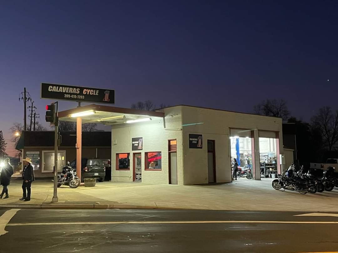 A motorcycle shop named Calaveras Cycle at dusk, with motorcycles outside, a sidewalk, and a few people walking.