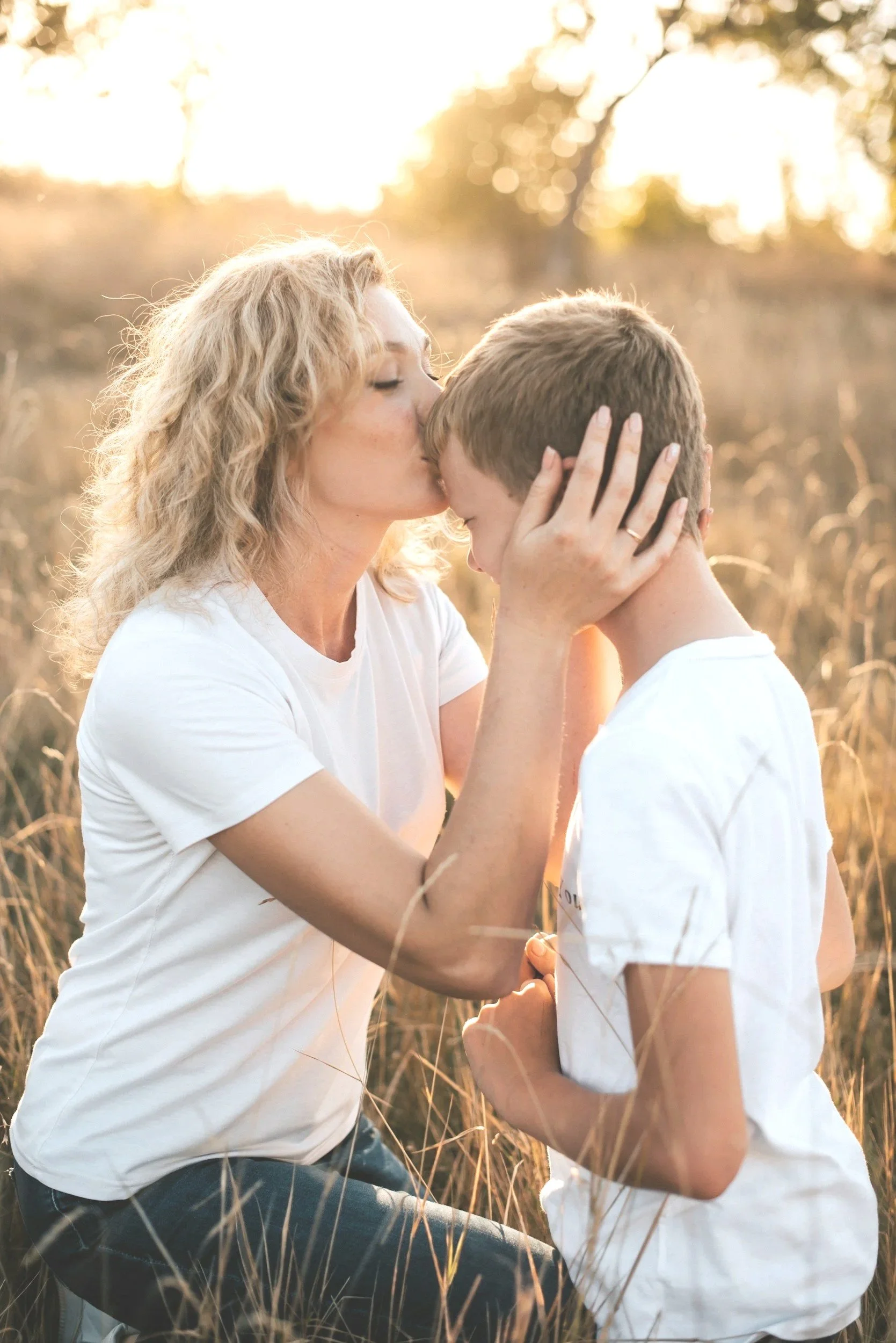 A woman with curly blonde hair kisses a young boy on the forehead in a field of tall grass at sunset.