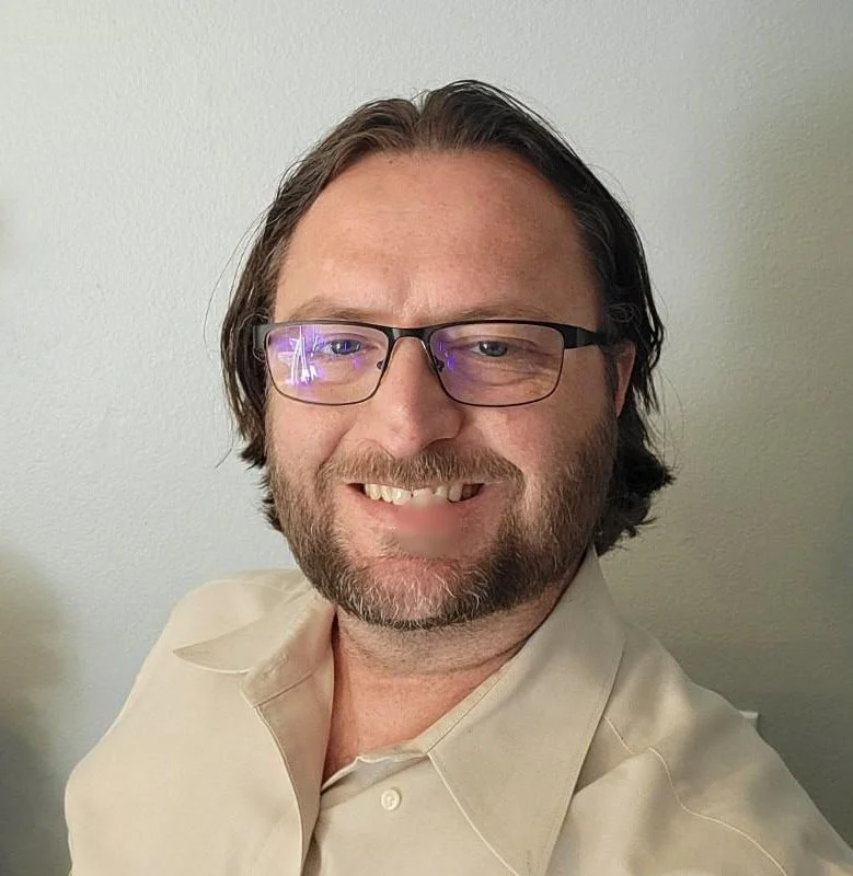 A man with long brown hair, glasses, and a beard smiling at the camera. He is wearing a light-colored collared shirt and standing against a plain wall.