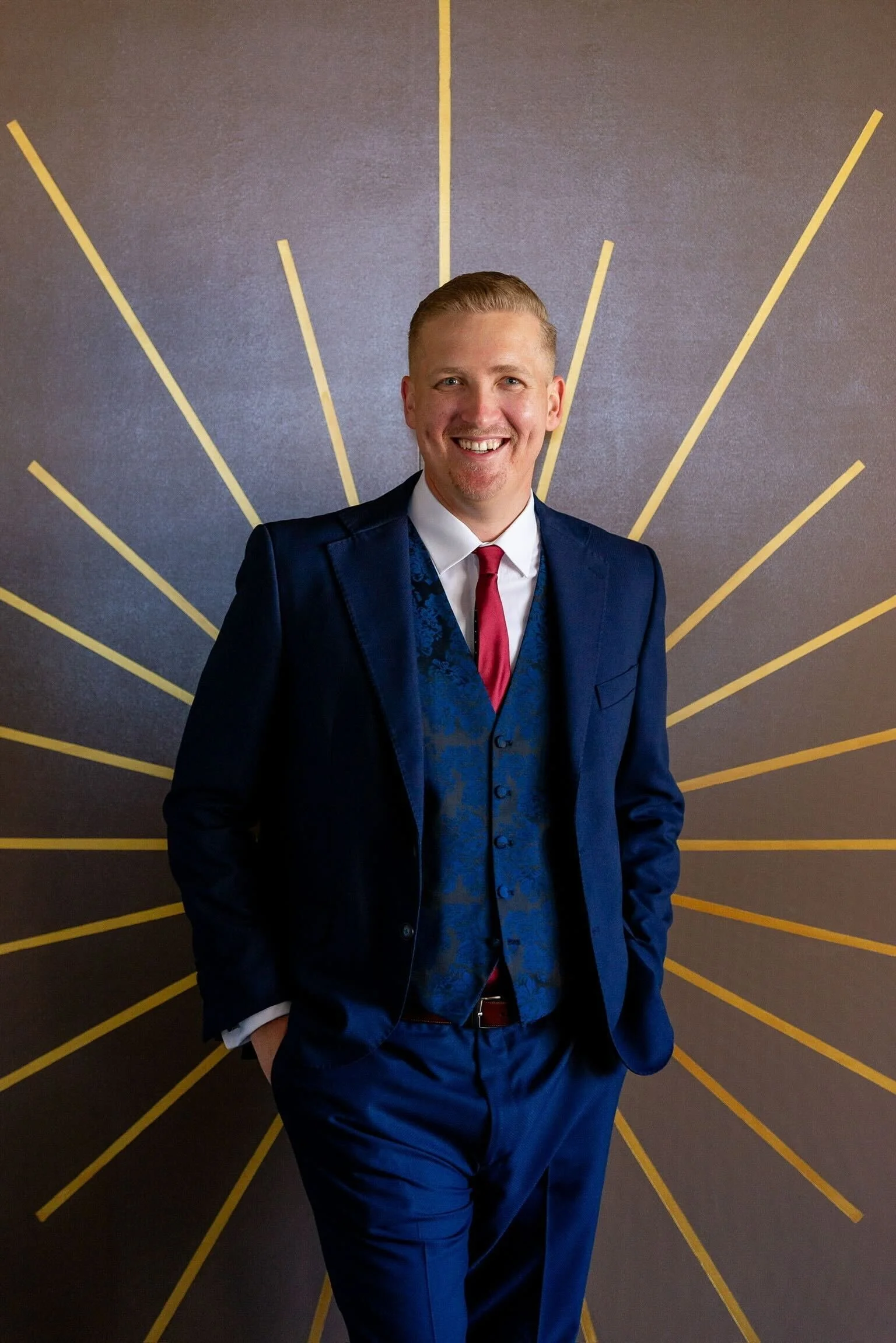 Smiling man in a blue suit, white shirt, red tie, and vest, standing in front of a decorative wall with radiating yellow lines.