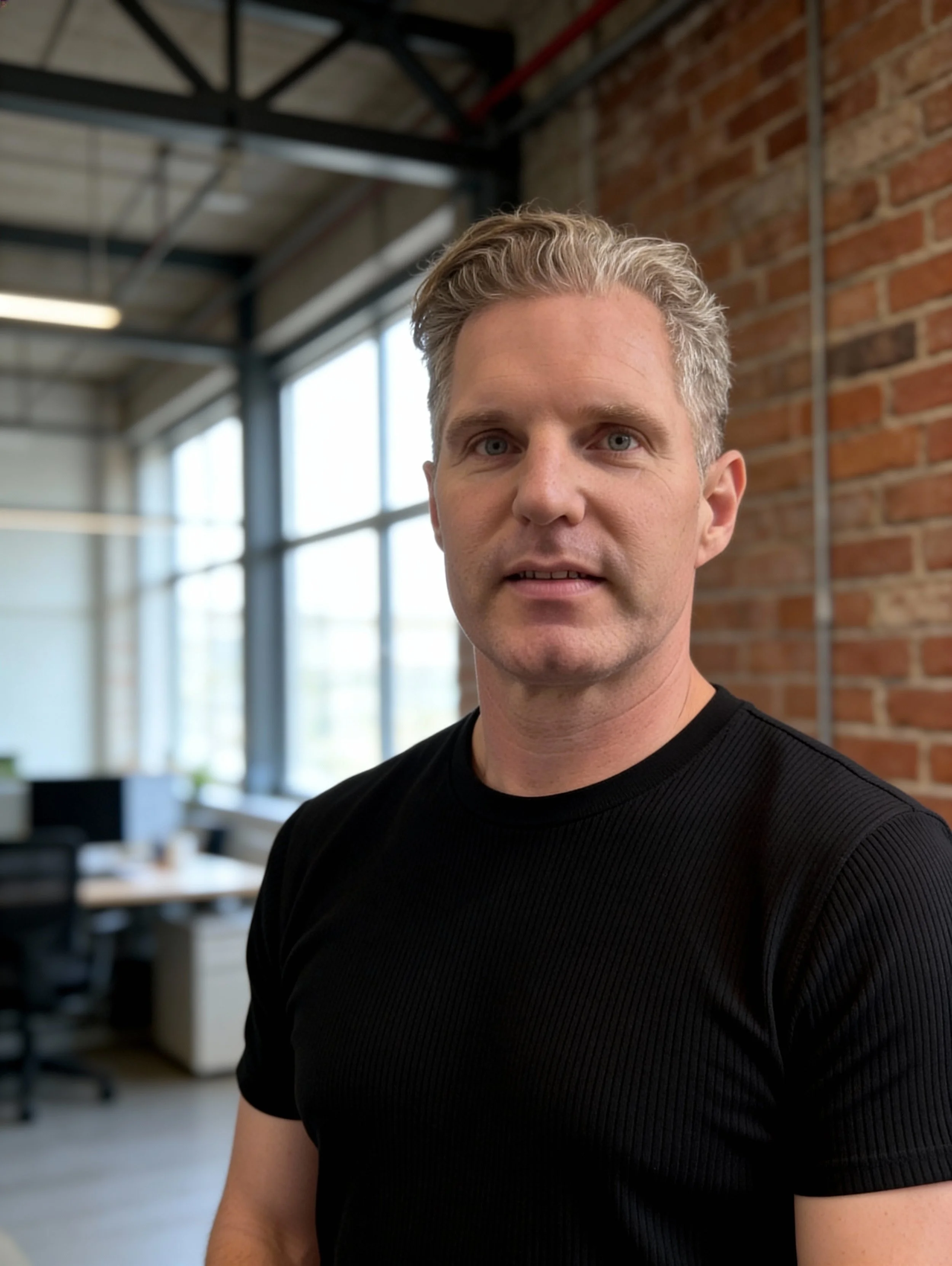 A man with light-colored hair and wearing a black shirt standing in an industrial-style office with large windows and exposed brick walls.