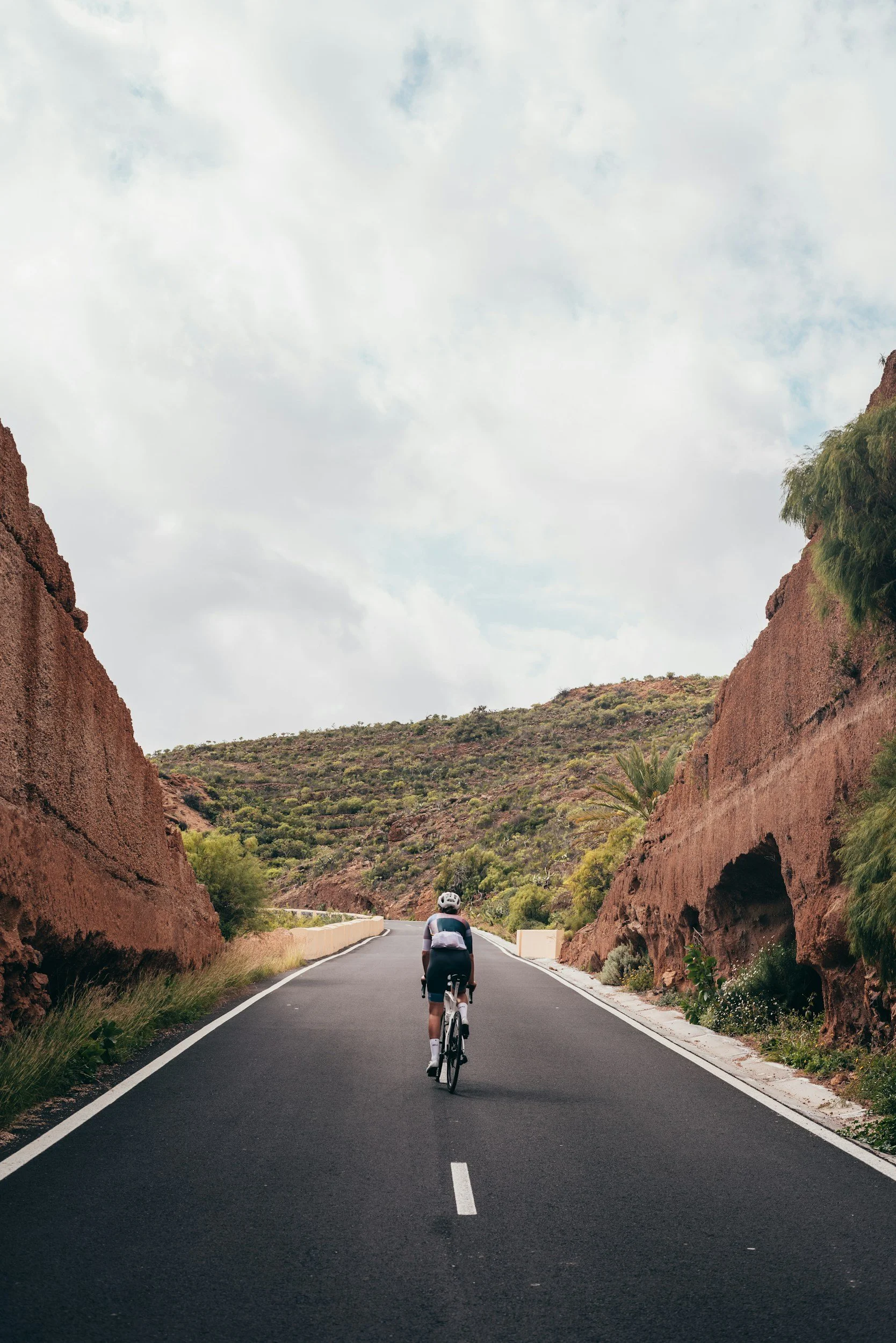 A cyclist riding on a paved road through a canyon with red rock walls on either side, green vegetation on the hills in the background, and a cloudy sky above.