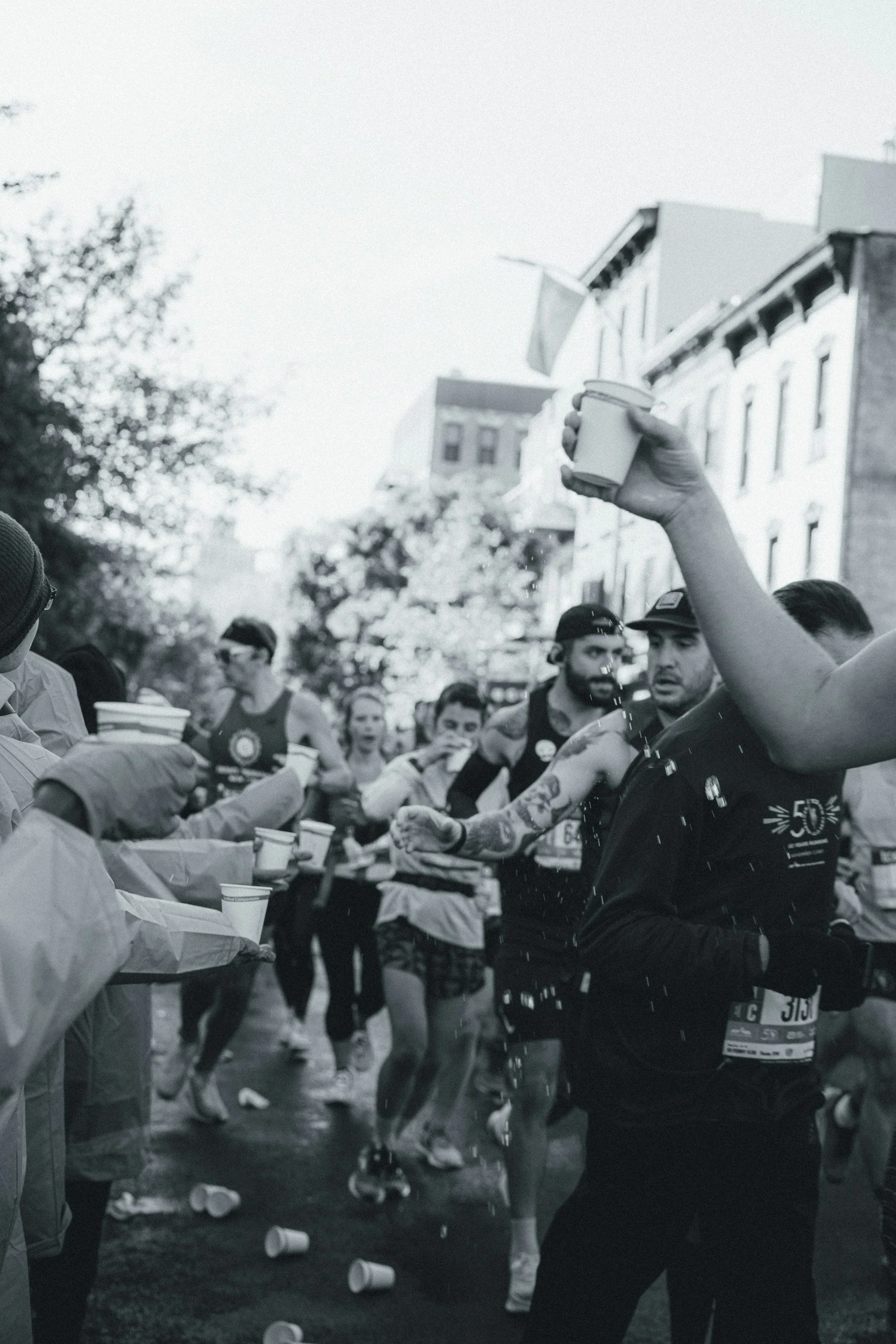 People participating in a running event, exiting cups on the ground, with some holding cups and water bottles, in a city street with buildings and trees.