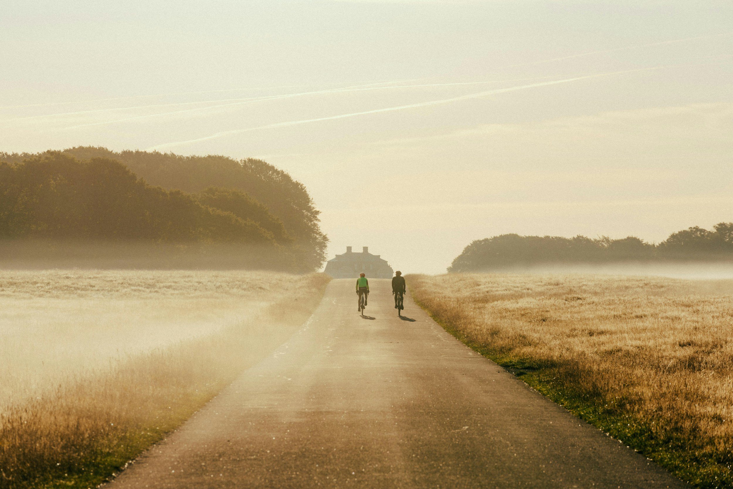 Two people cycling on a narrow country road surrounded by fields and trees in early morning light.
