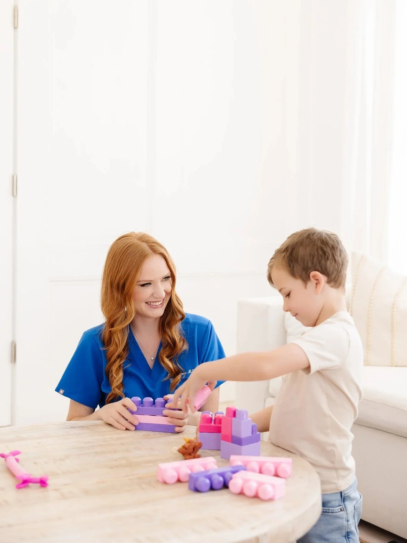 A young woman with long red hair, wearing a blue nurse uniform, is smiling at a young boy with light brown hair, playing with pink and purple toy blocks at a wooden table in a bright, white room.