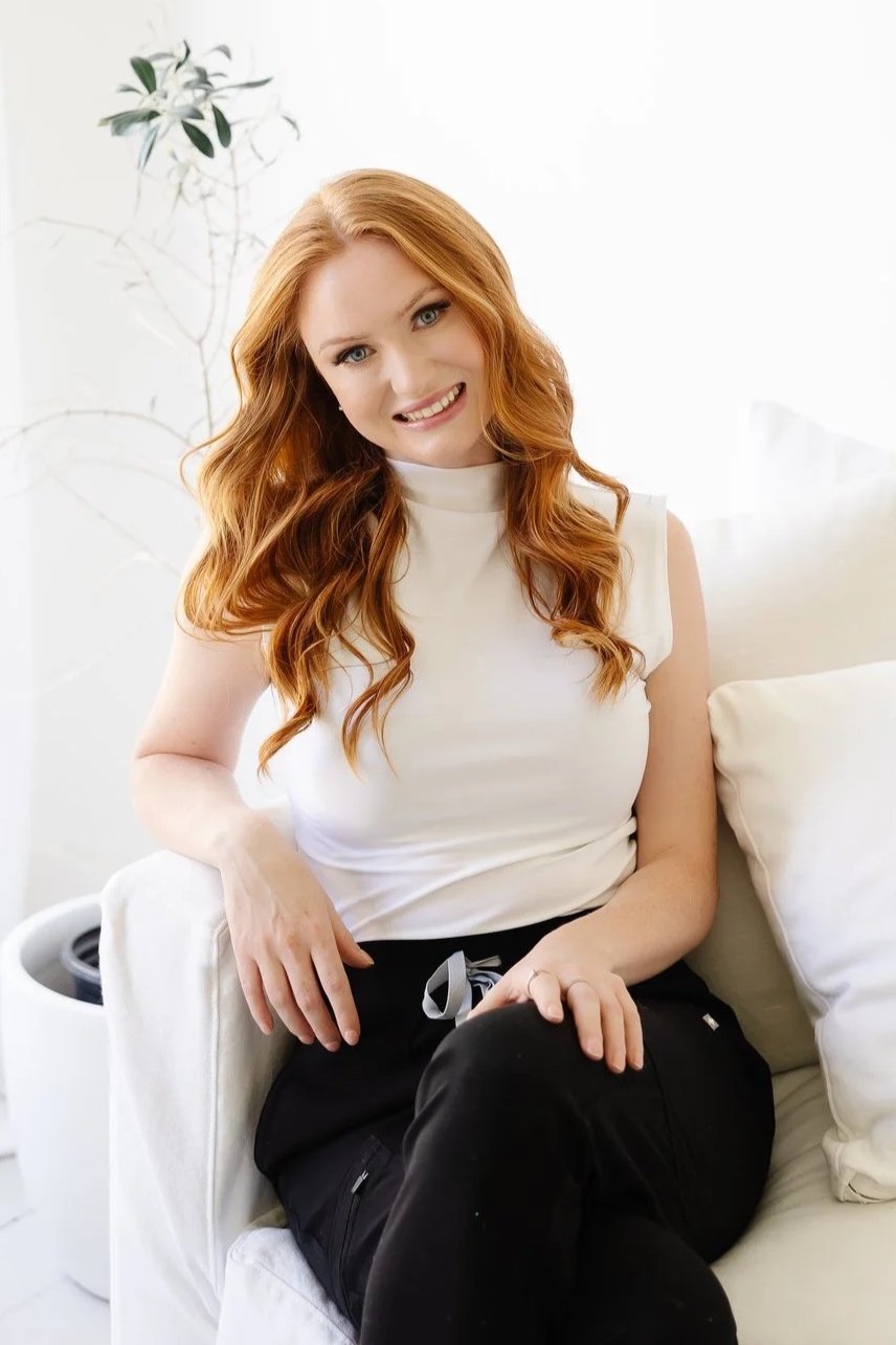 A young woman with long red hair, wearing a white sleeveless top and black pants, sitting on a white couch in a bright room, smiling at the camera.