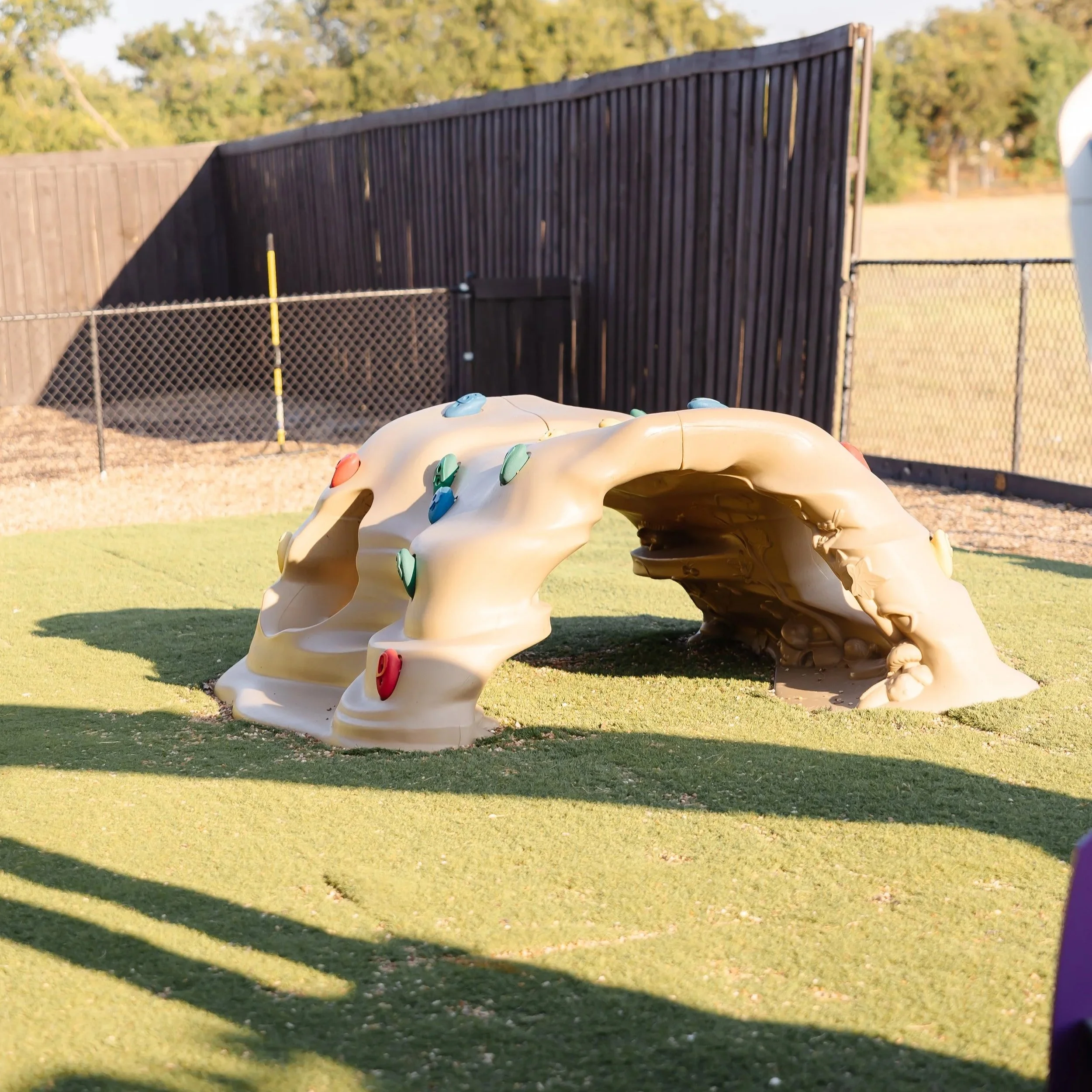 Plastic climbing structure shaped like a rock with colorful handholds on a grassy area inside a fenced playground.
