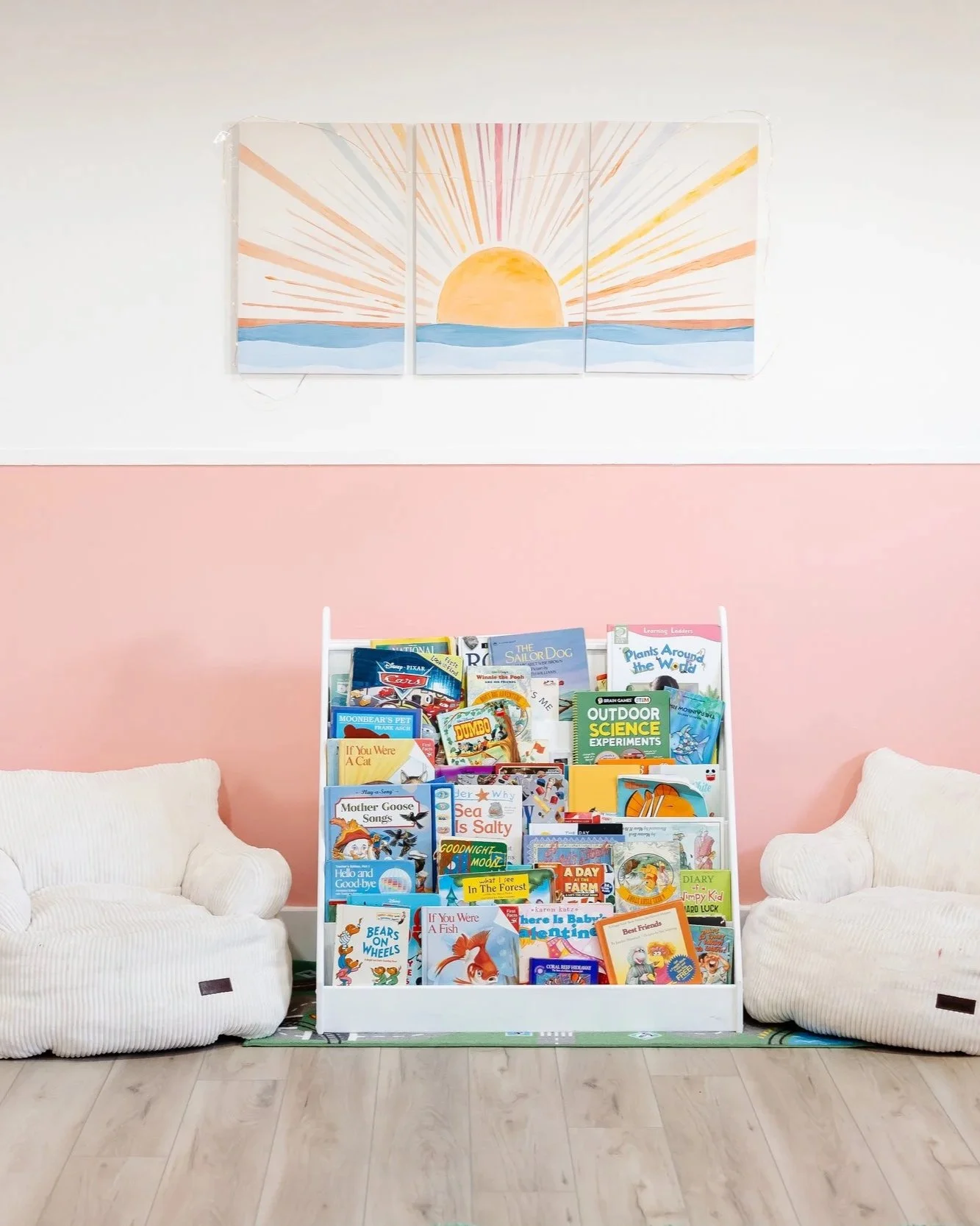 Children's bookshelf filled with colorful books flanked by two large white plush armchairs in a room with pink and white walls and a sun painting above.