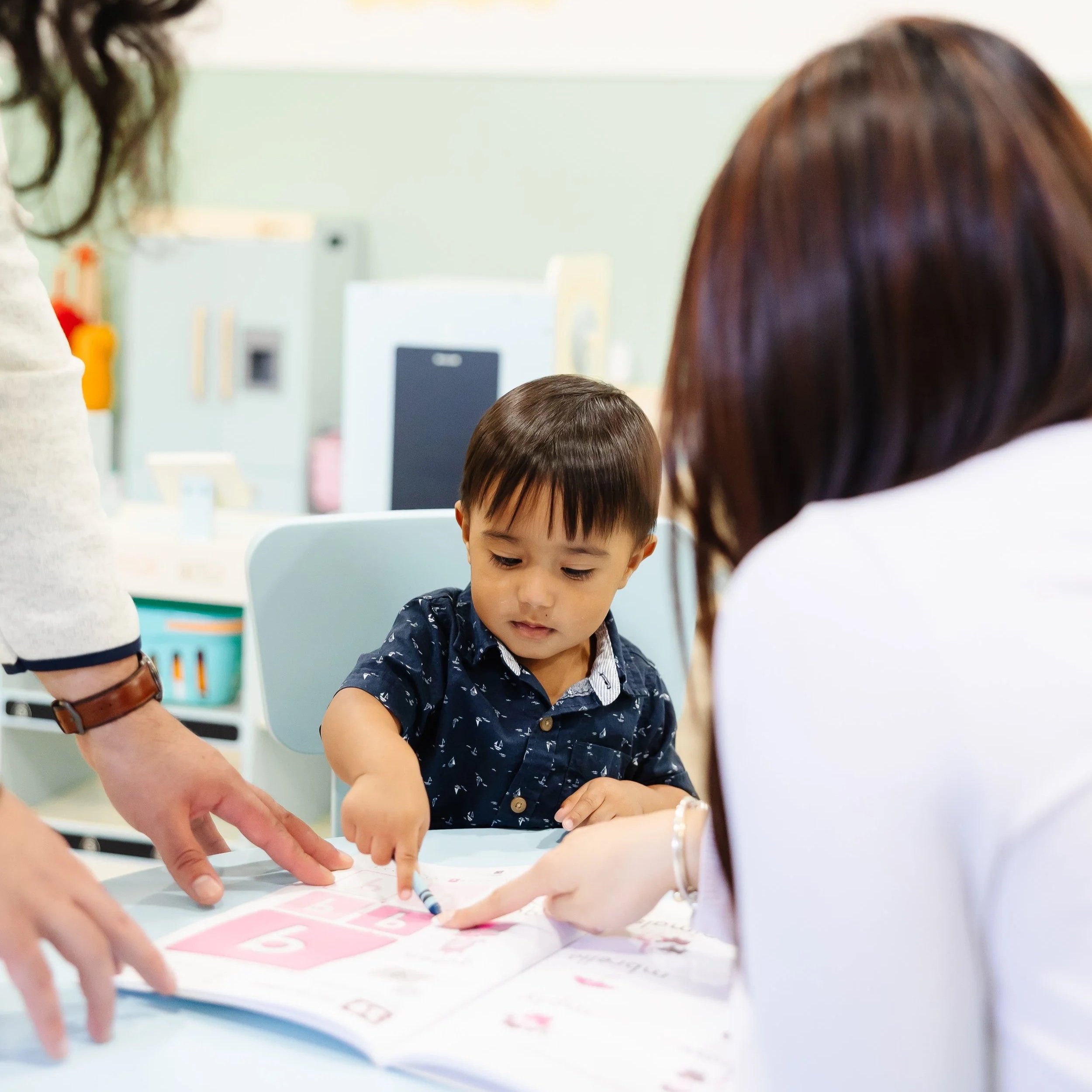A young boy sitting at a table with two adults, pointing at a colorful educational book with pictures and letters, in a classroom or therapy setting.