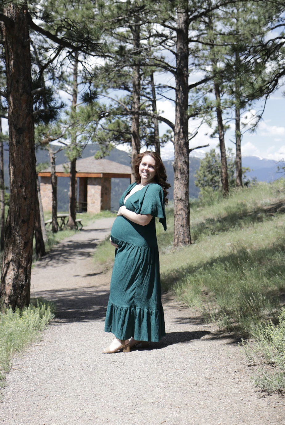 A pregnant woman with red hair wearing a teal dress and beige heels standing on a dirt trail in a wooded park with pine trees, mountains, and a small stone pavilion in the background.