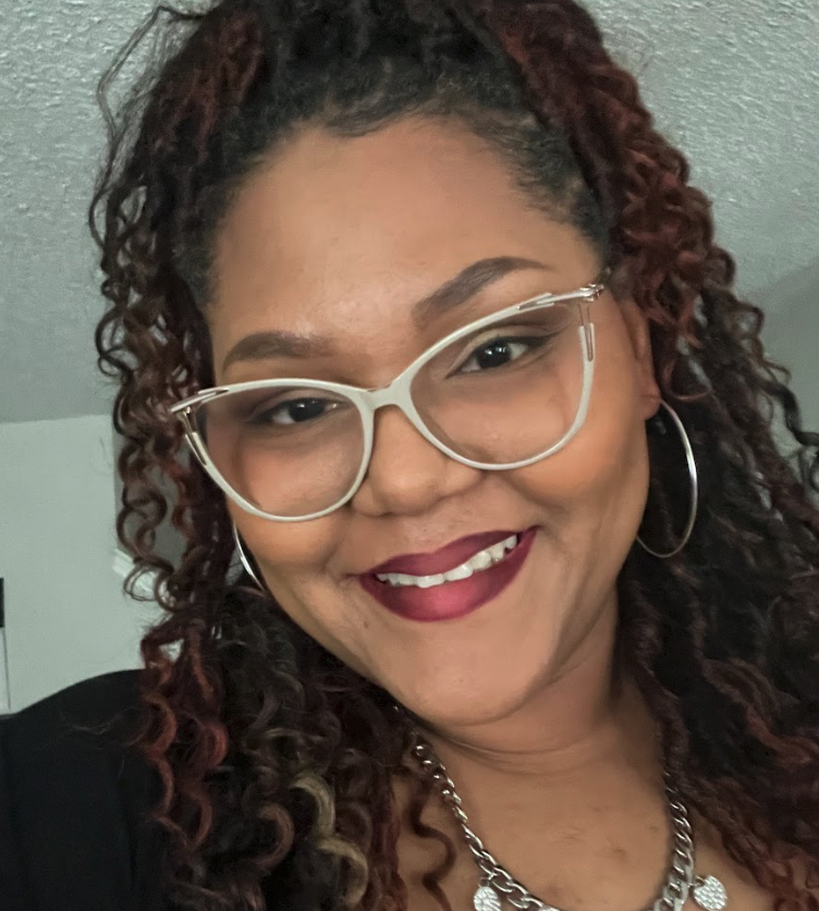 Smiling woman with curly hair, wearing large glasses, hoop earrings, and a chain necklace.