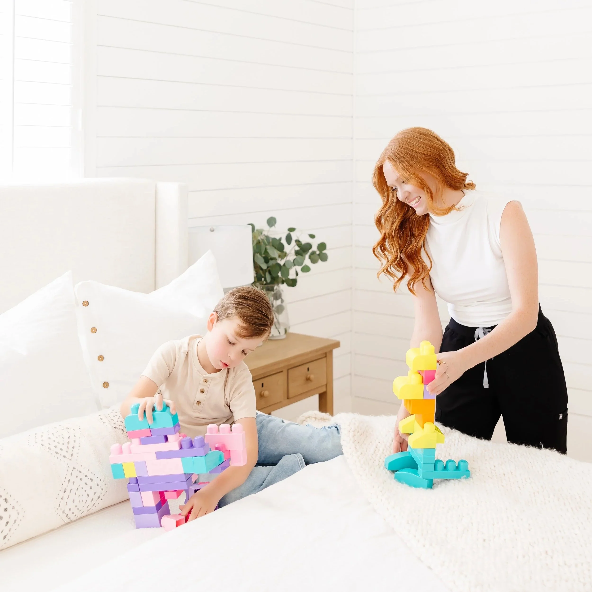 A woman and a young boy playing with colorful stacking blocks on a bed in a bright, white bedroom.