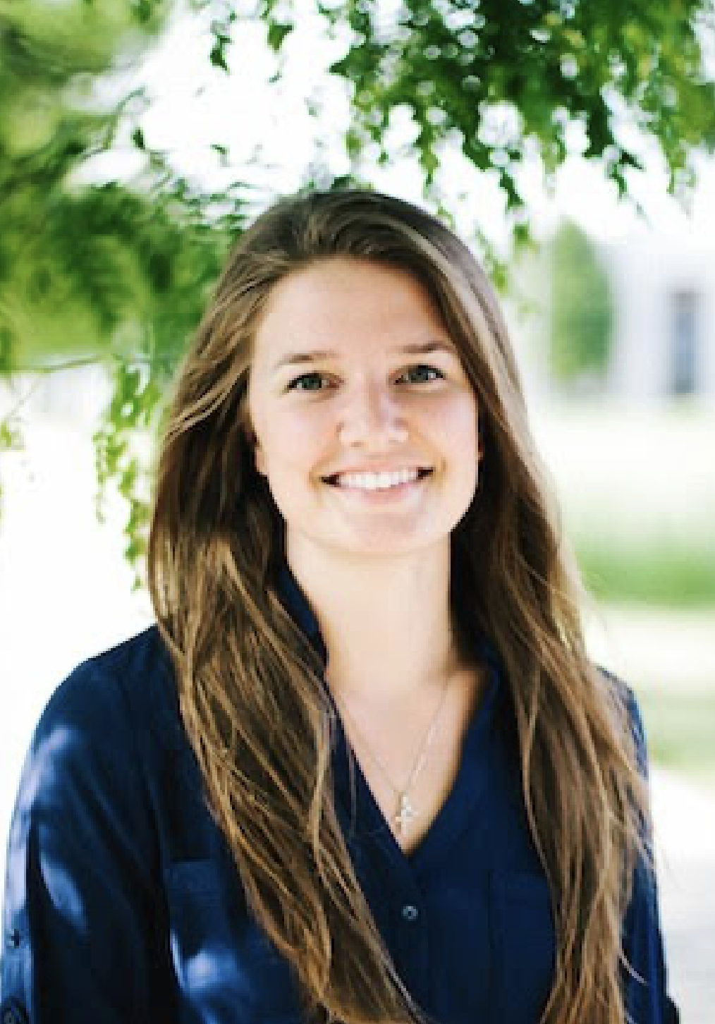 A young woman with long brown hair smiling outdoors, dressed in a dark blue blazer, standing under a tree with green leaves.