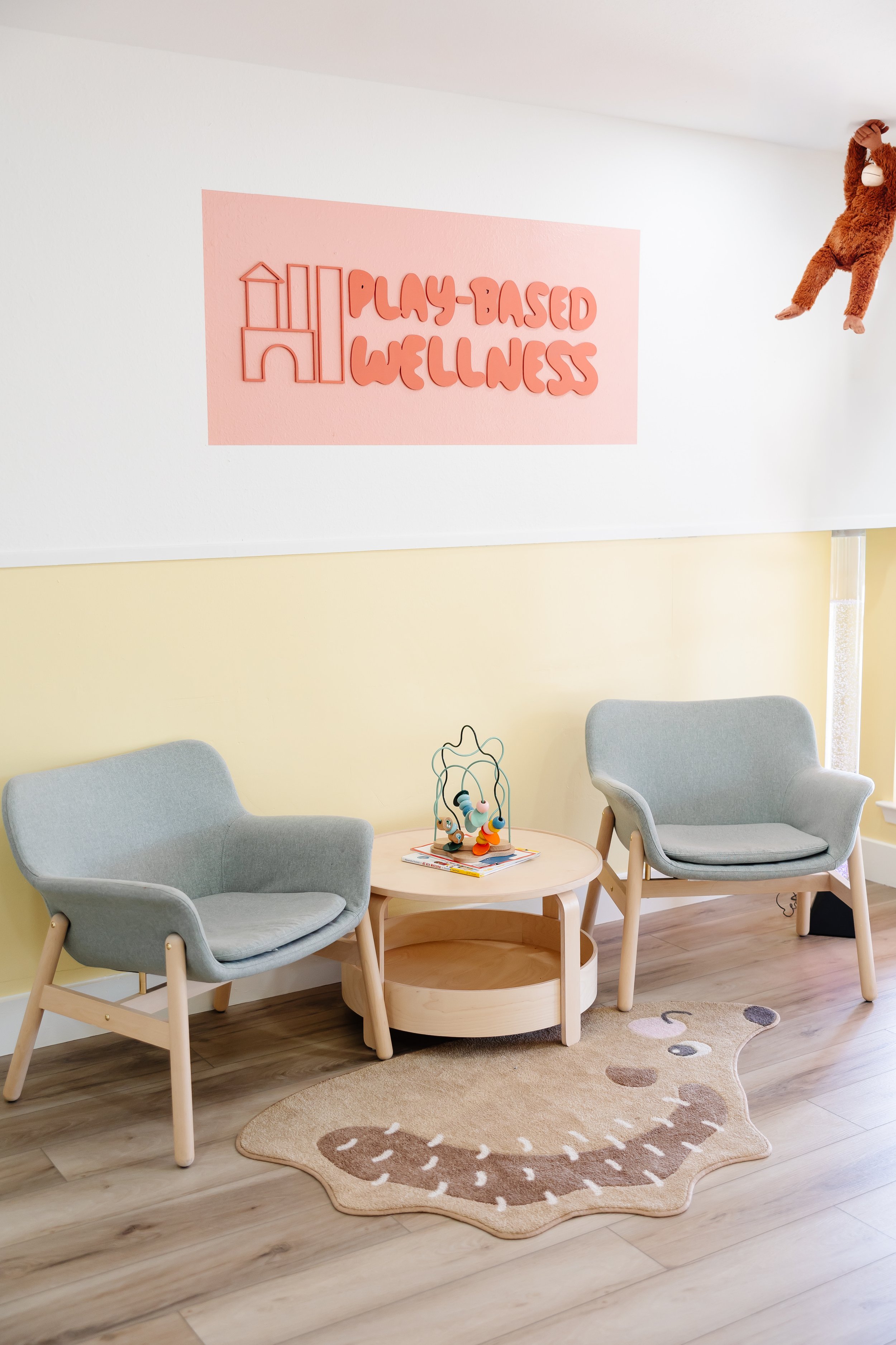 A waiting area with two light green armchairs, a round wooden coffee table, and a decorative rug with a smiling animal face. There is a pink sign on the wall that says "Play-Based Wellness" and a hanging stuffed monkey toy.